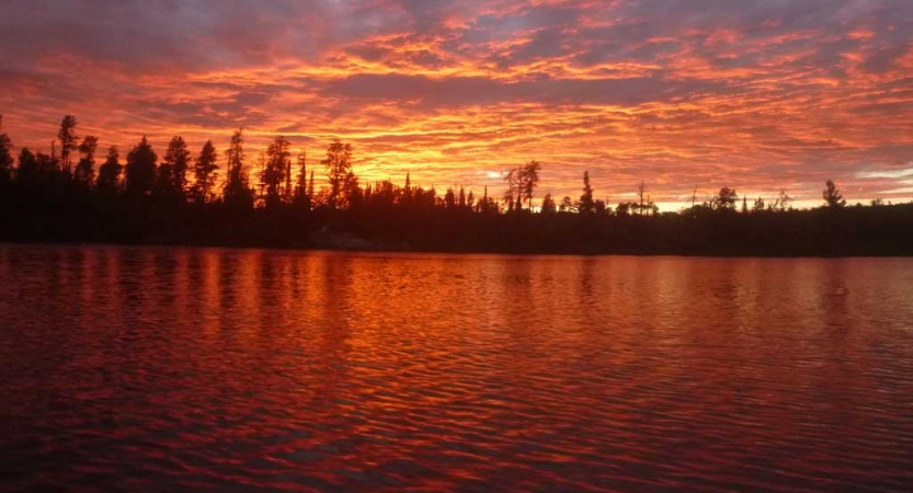 The sky appears in shades of red, orange and purple, reflected in the lake below, between a line of trees. 